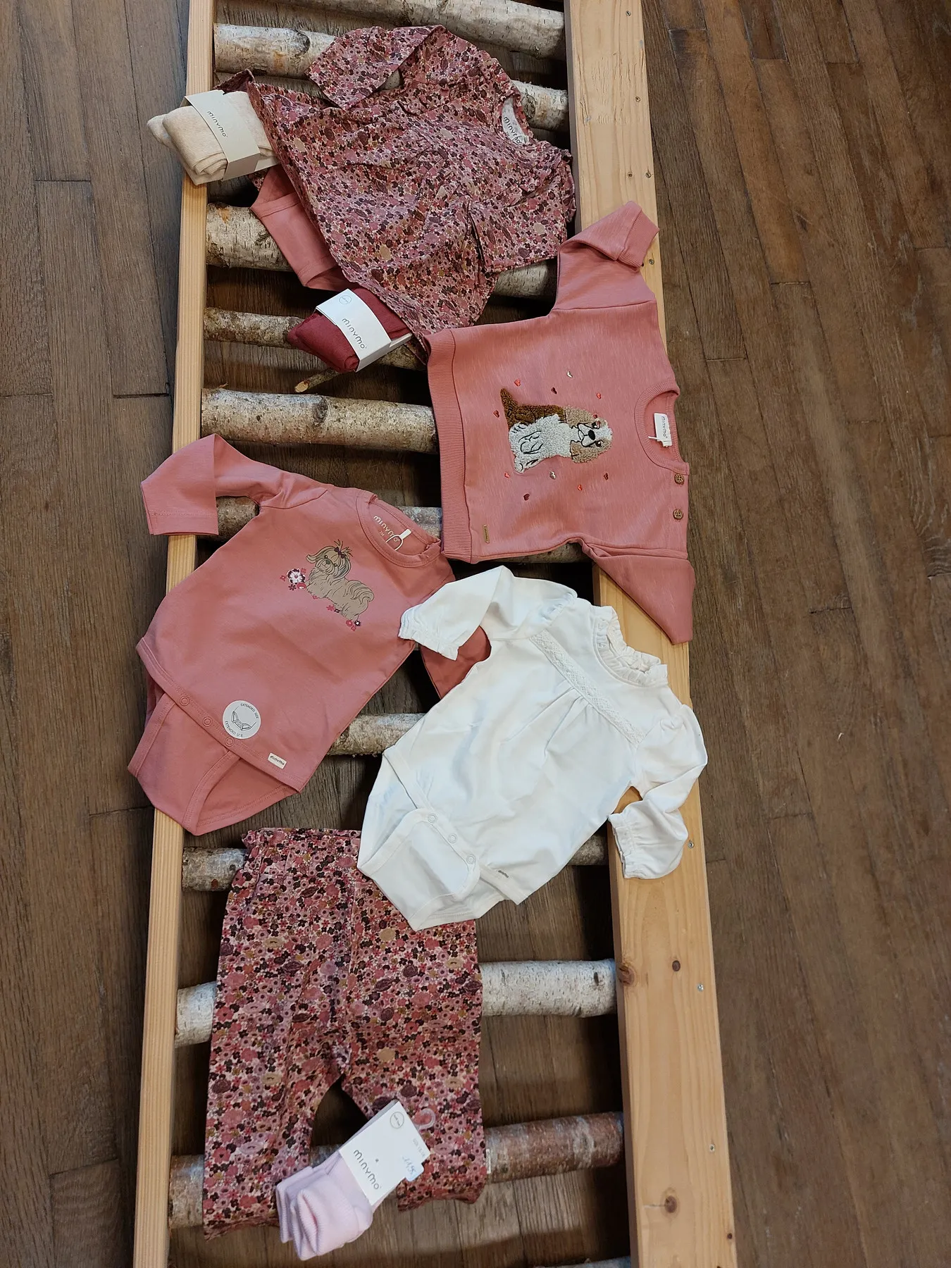 Des tenues de bébé en tons roses disposées sur un présentoir en rondins de bouleau.
