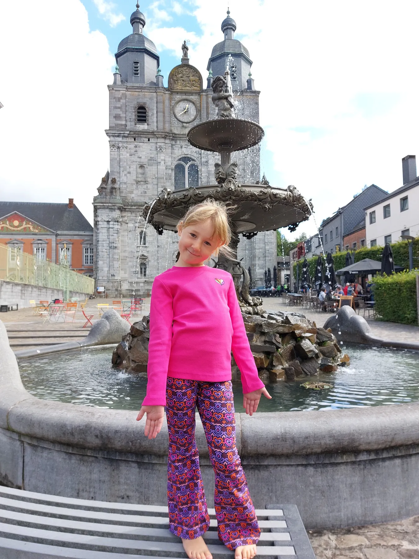 Une petite fille en tenue de la boutique pose devant la fontaine et la basilique de Saint-Hubert.