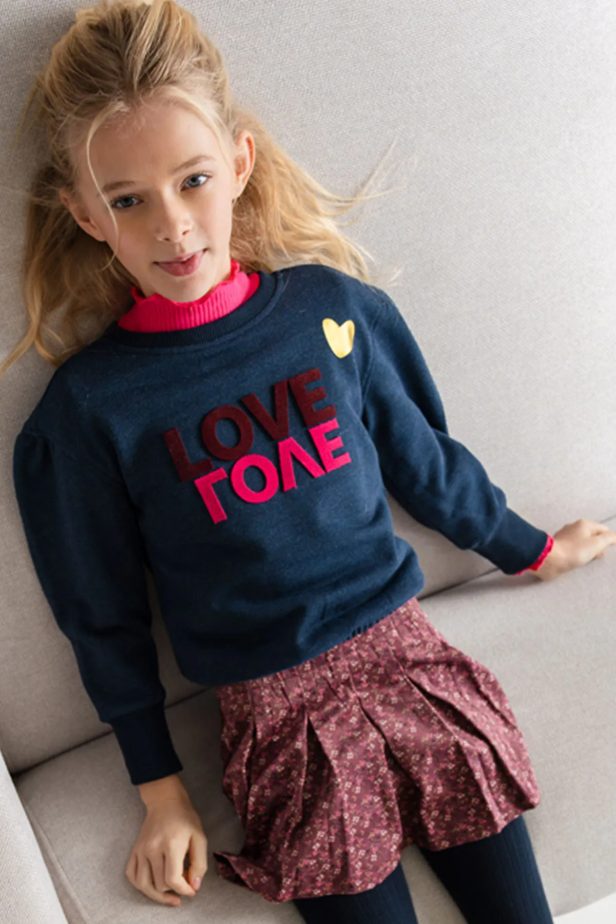 Une fillette porte un pull marine à inscription et une jupe à fleurs bordeaux.